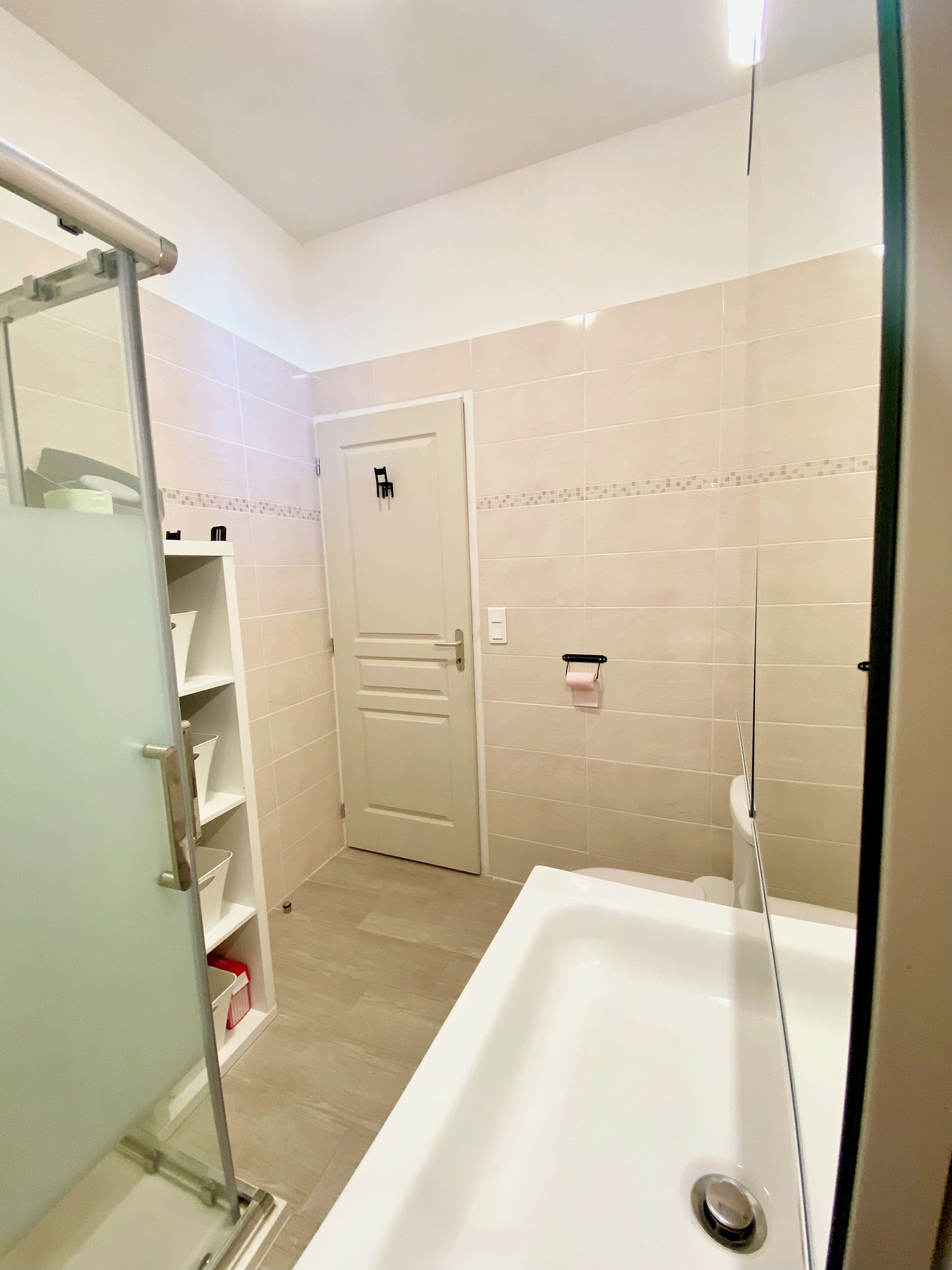 Un baño con bañera, zona de ducha e inodoro. Hay estantes con artículos de aseo y un perchero para toallas.