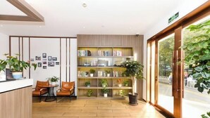 A reception area with a wooden reception desk, leather chairs, a bookshelf with books and plants, and a large window with a view of the outdoors. - Jizhu Auto Business Hotel (Shanghai)