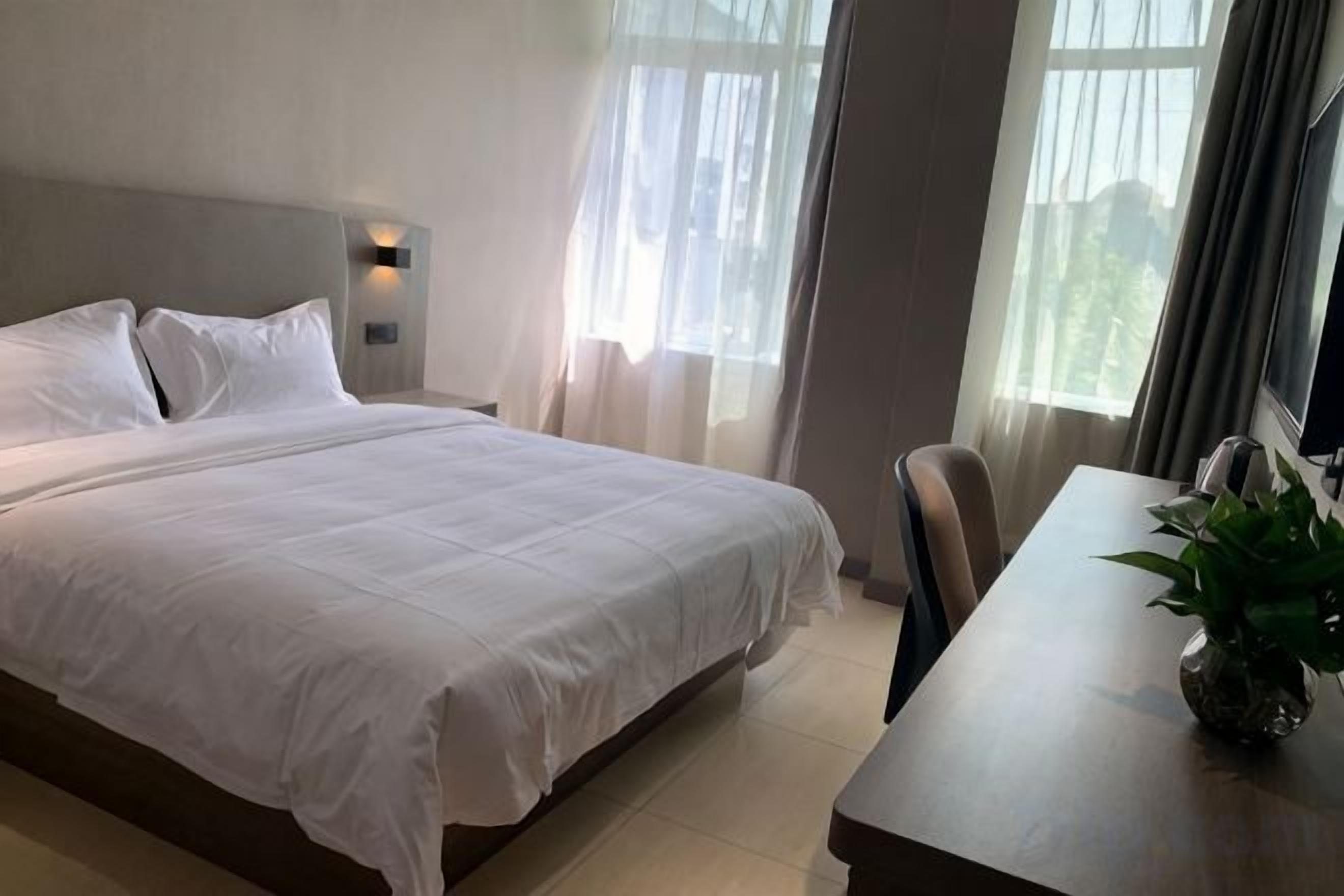 Een hotelkamer met een bed, nachtkastje met een plant, een stoel en een raam met gordijnen.