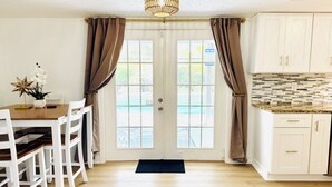 A dining area with a wooden table and white chairs, a kitchen with white cabinets and a tiled backsplash, and a door with brown curtains. - Tampa Luxury 5BR Estate with Pool + Golf Views (Tampa)