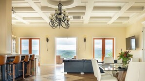 A spacious living area with a large sofa, a dining table with chairs, a kitchen area with bar stools, and a view of the outdoors through large windows.