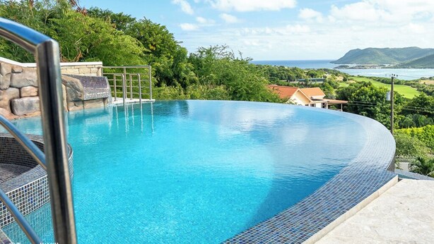 A clear blue infinity pool with a stone and metal deck, surrounded by lush greenery and a view of the ocean and hills.