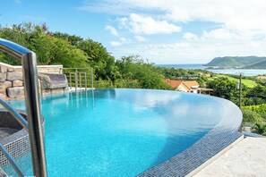 A clear blue infinity pool with a stone and metal deck, surrounded by lush greenery and a view of the ocean and hills.