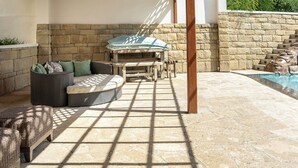 A sunlit patio with stone flooring, a wicker sofa, a wooden table, and a stone wall.