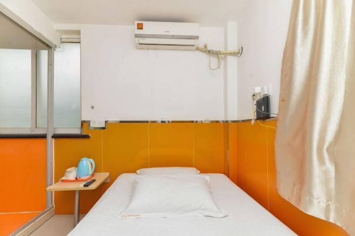 A single bed with a white mattress and pillows, an air conditioning unit mounted on the wall, a small table with a kettle and a remote control, and a window with blinds.