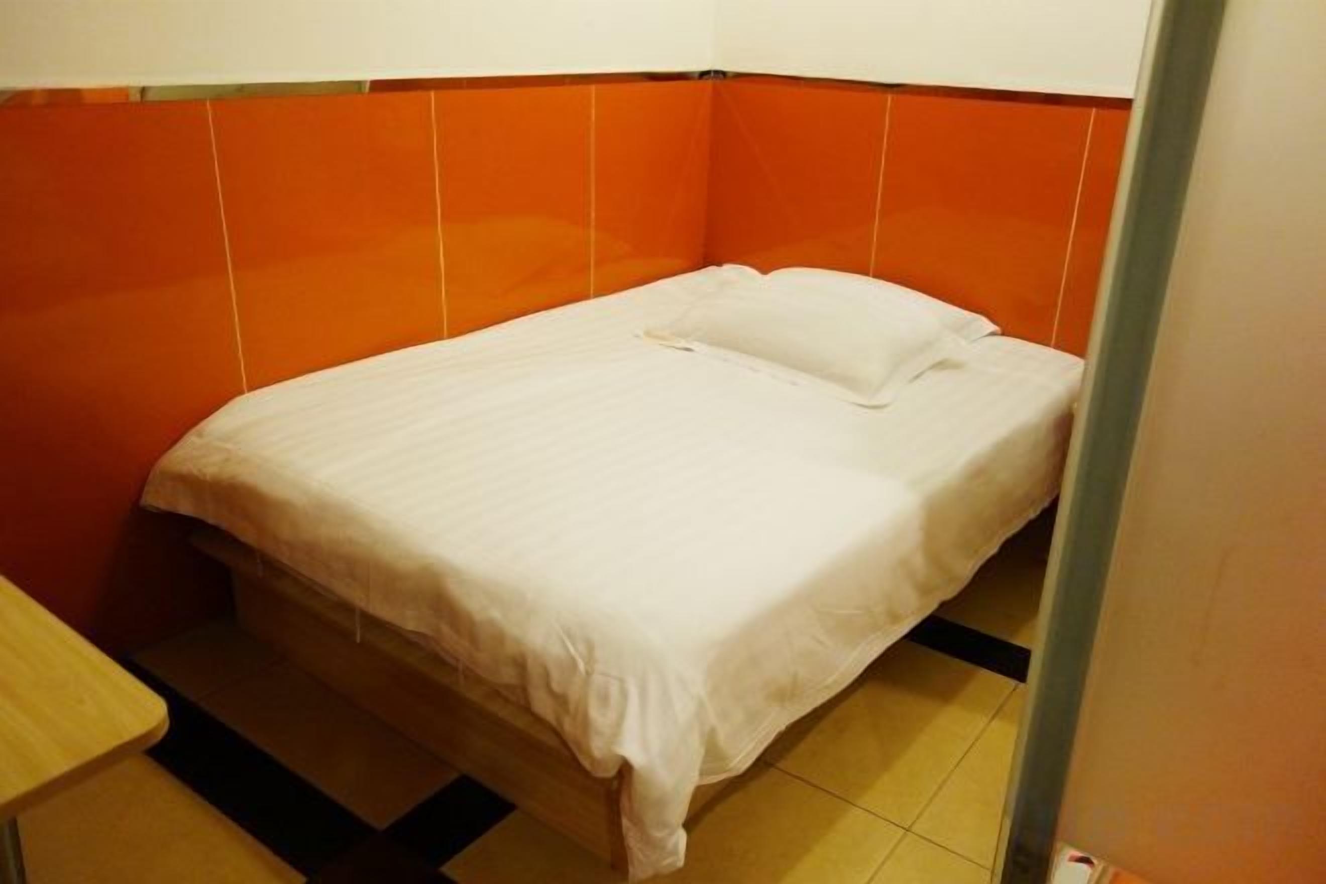 A single bed with a white mattress and pillow in a room with orange walls and a tiled floor.