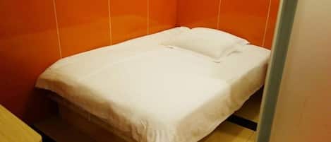 Una cama individual con colchón y almohada blancos en una habitación con paredes anaranjadas y suelo de baldosas.