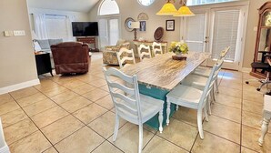 A spacious living room with a large dining table, white chairs, a brown leather sofa, and a television.