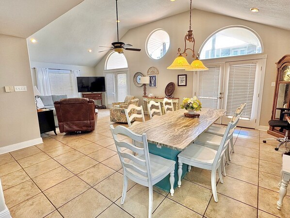 A spacious living room with a large dining table, white chairs, a brown leather sofa, and a television.