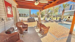 A covered patio area with wicker furniture, a ceiling fan, and a view of a pool and buildings in the background.