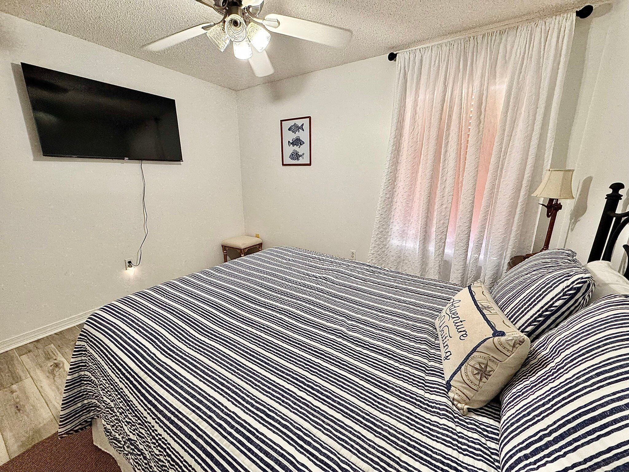 A bedroom with a bed, a ceiling fan, a TV mounted on the wall, a small bedside table, and a window with curtains.