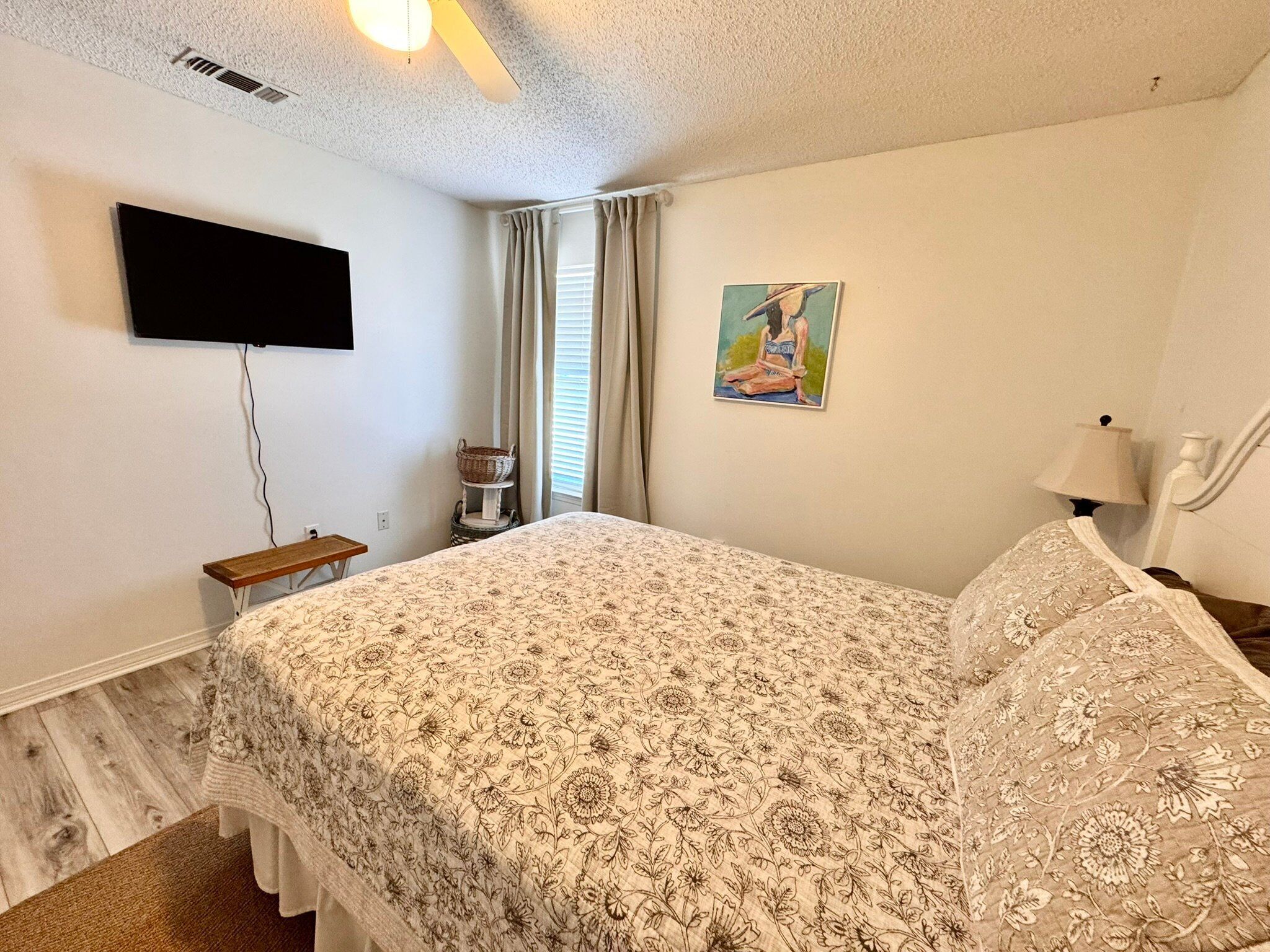 A bedroom with a bed, a flat-screen TV, a painting, a bedside lamp, and a small table.