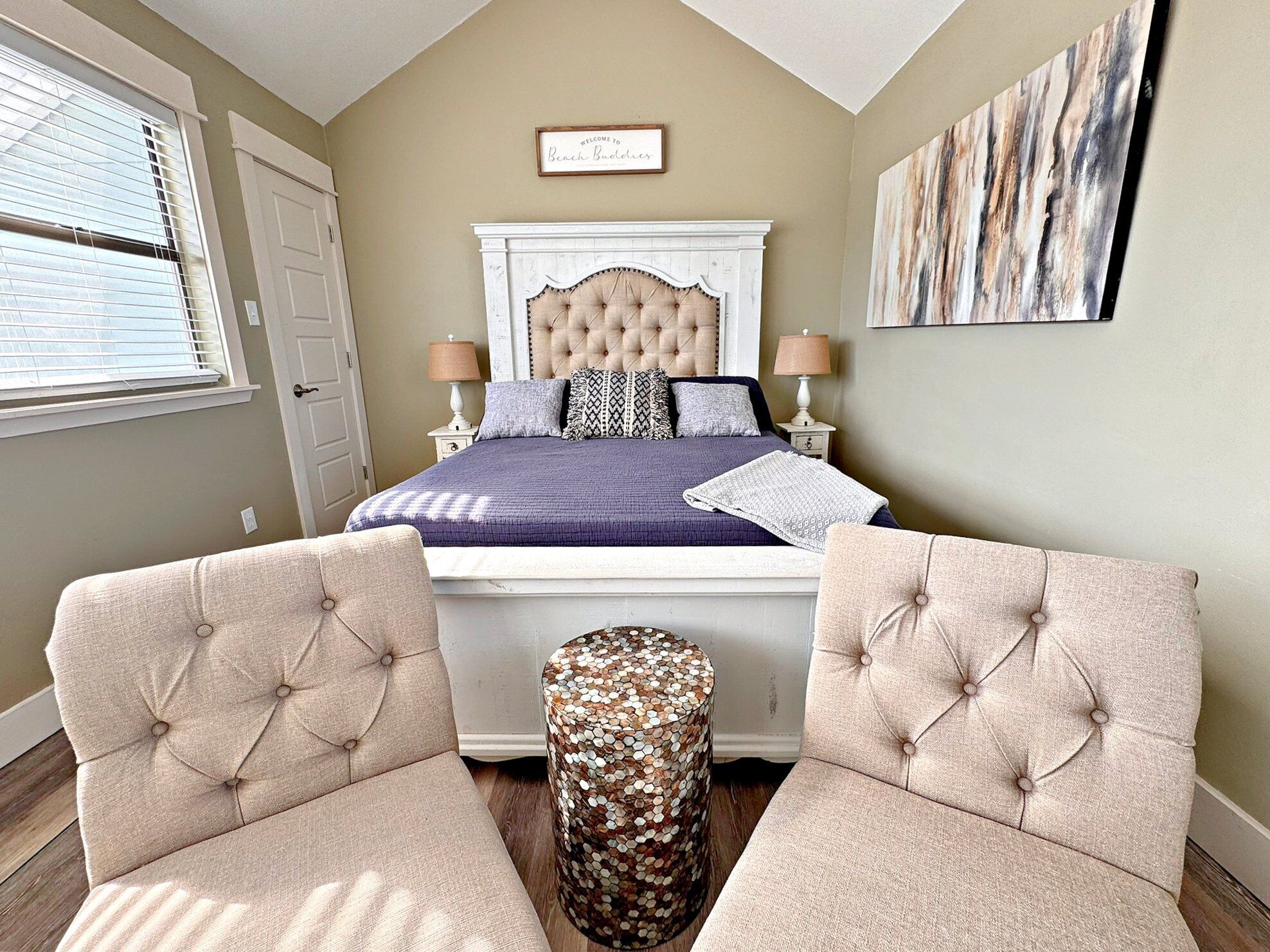 A bedroom with a large bed, two armchairs, a side table, and a decorative wall piece.