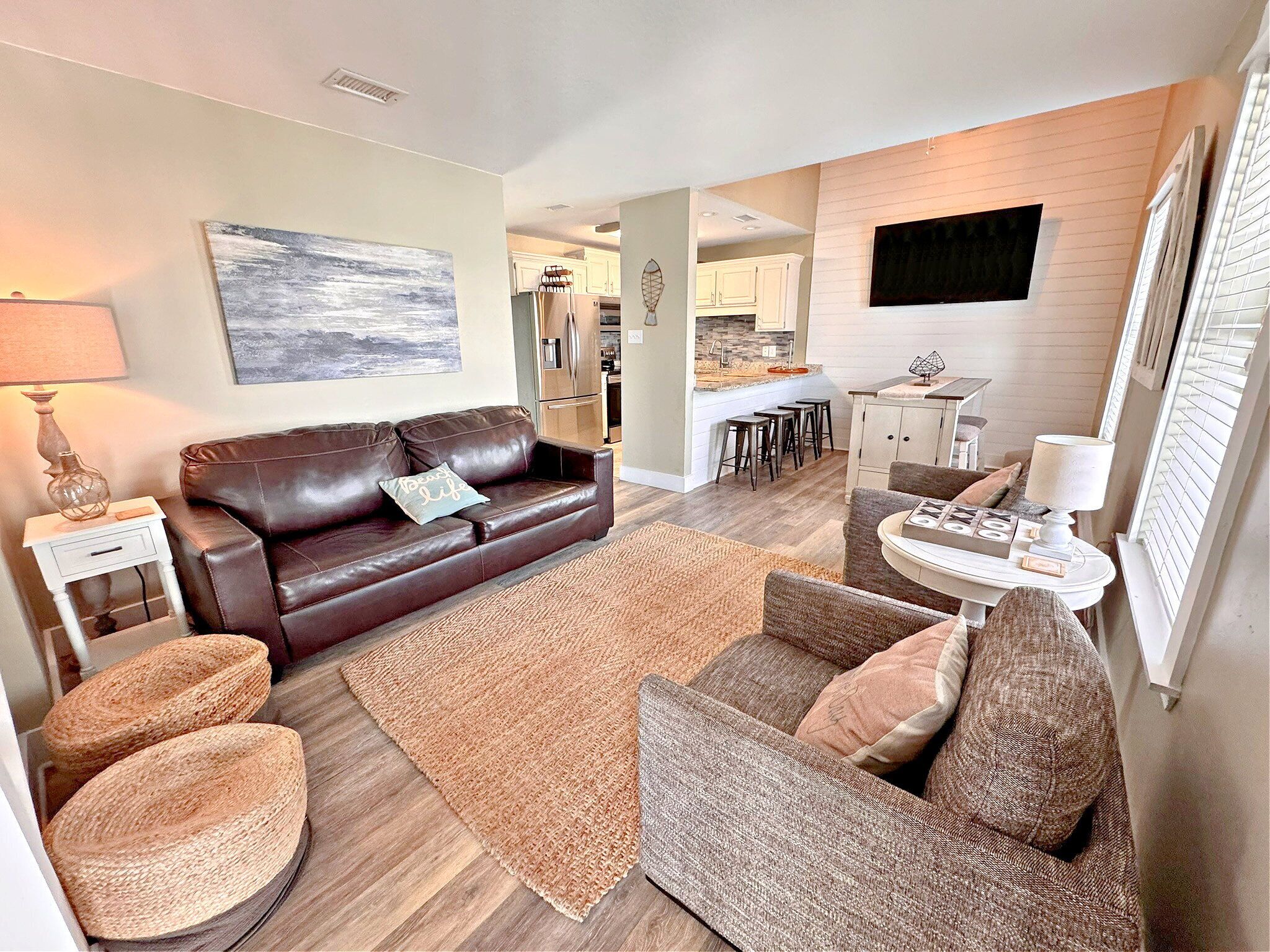 A modern living room with a brown leather sofa, a round wicker chair, and a small white table. A kitchen is visible in the background with white cabinets and a black countertop.