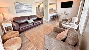 A modern living room with a brown leather sofa, a round wicker chair, and a small white table. A kitchen is visible in the background with white cabinets and a black countertop. - Ocean Reef 504 (Gulf Shores)