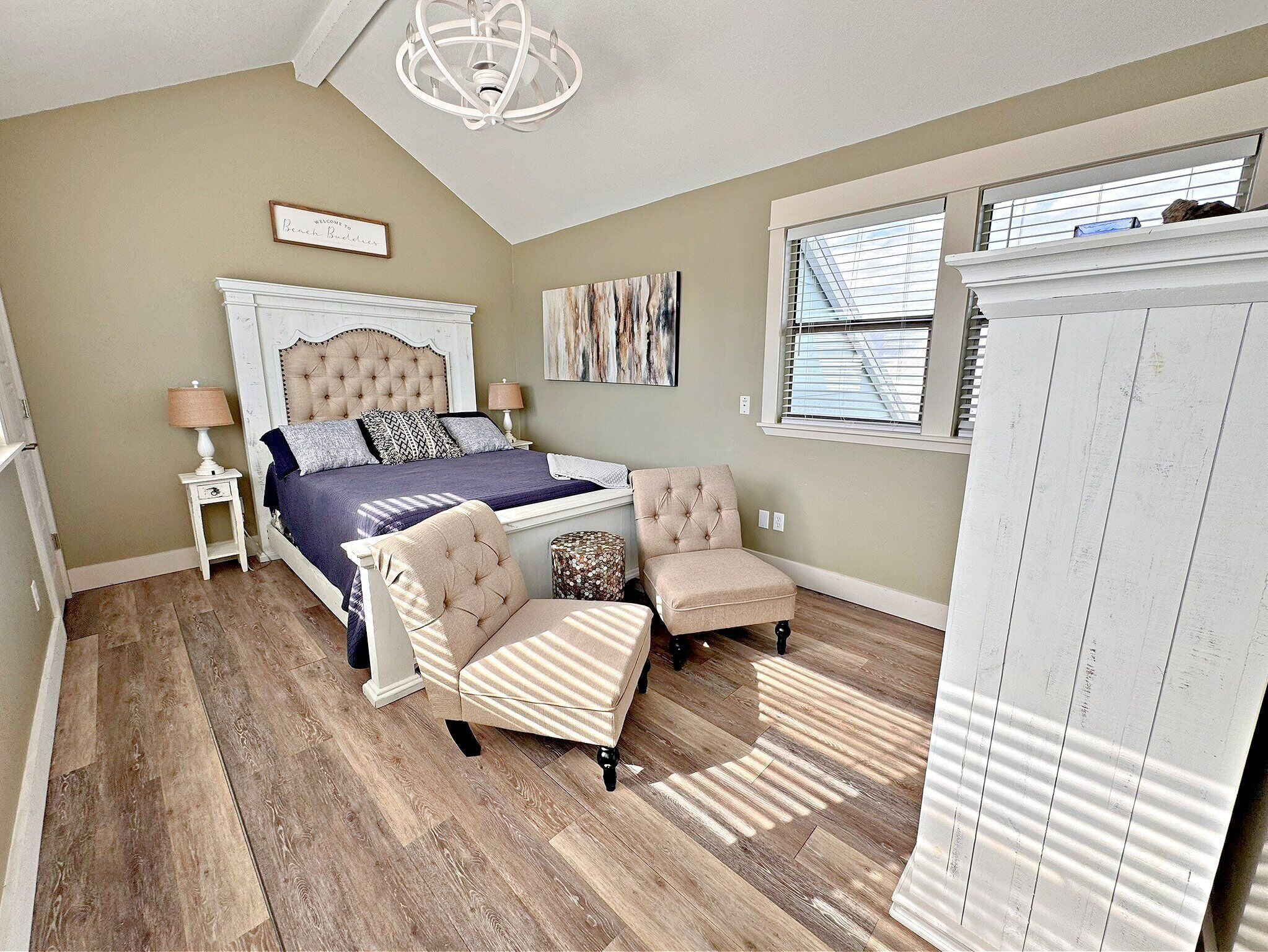 A bedroom with a large bed, two armchairs, a nightstand, a dresser, a ceiling fan, and a window with blinds.