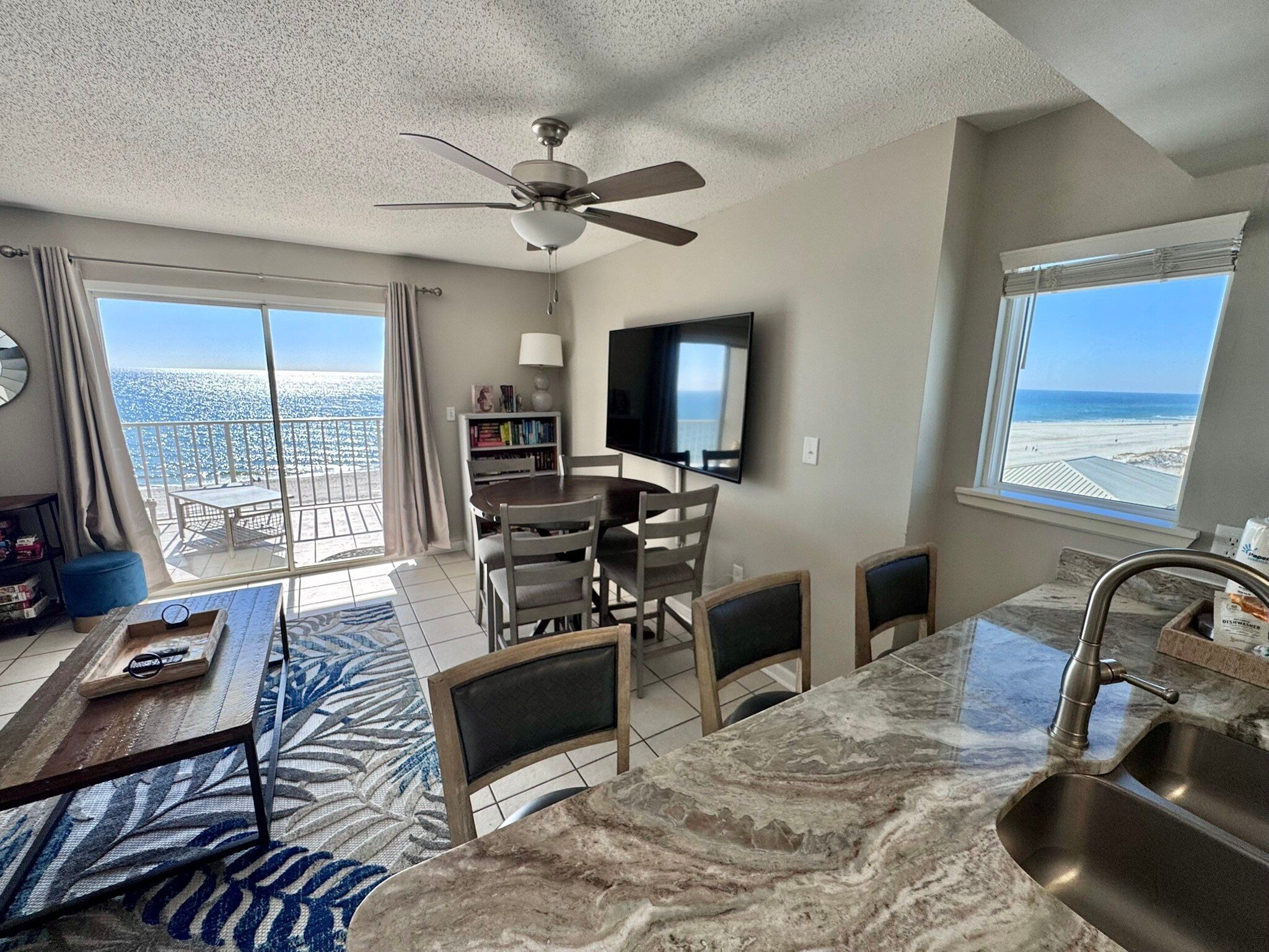 A living room with a dining area, a kitchen with a sink, a ceiling fan, and a TV. There is a balcony with a view of the beach.