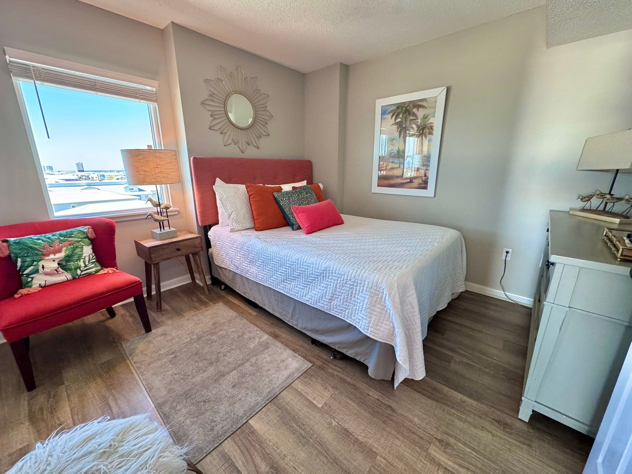 A bedroom with a bed, a nightstand, a red chair, a window with blinds, a wall-mounted mirror, and a framed picture.