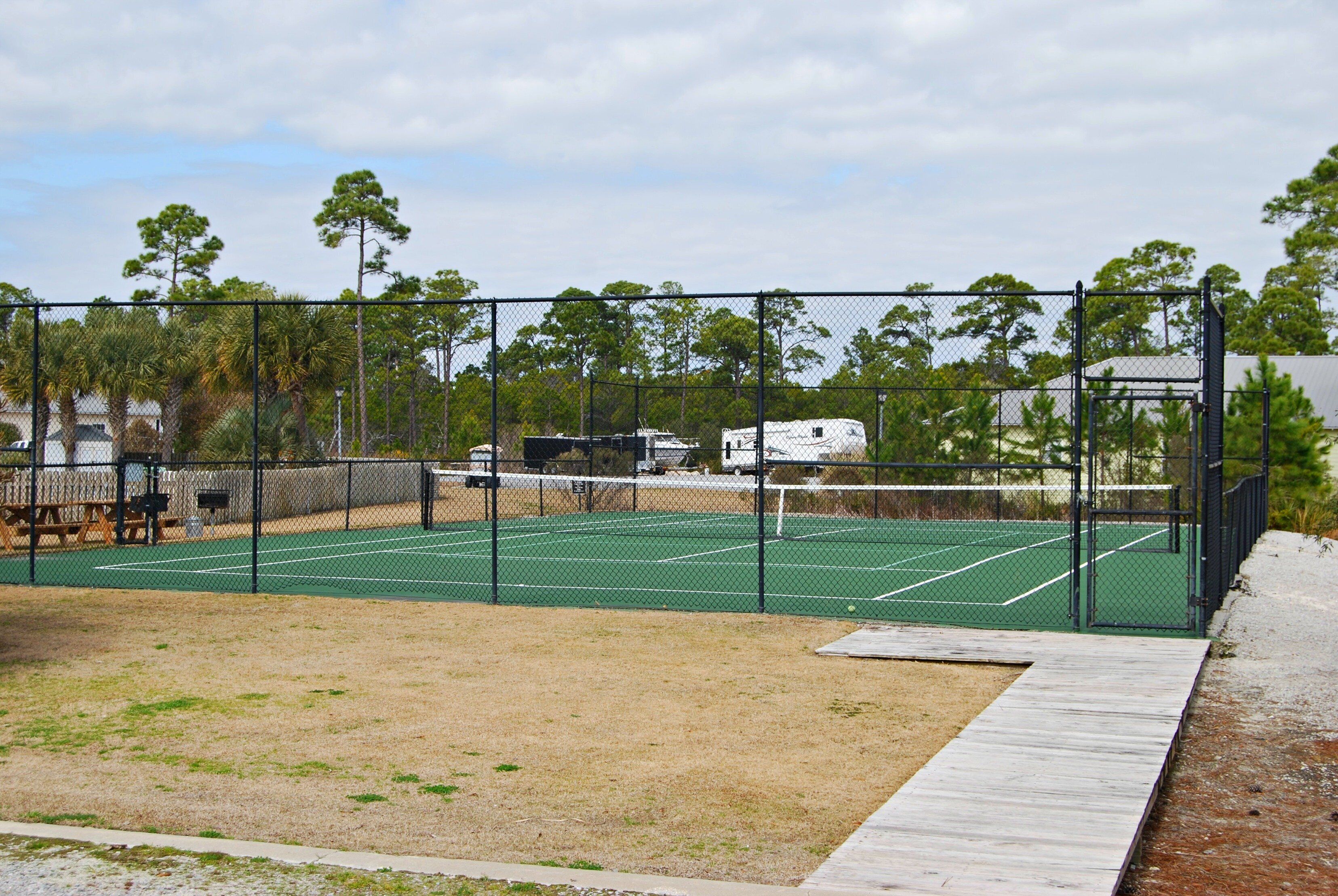 Un court de tennis doté d’une terrasse en bois, entouré d’une clôture grillagée, avec des arbres en arrière-plan.