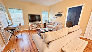 A living room with a sofa, a coffee table, a TV on a stand, and a door with a fish decoration. - The Rookery 202 (Gulf Shores)