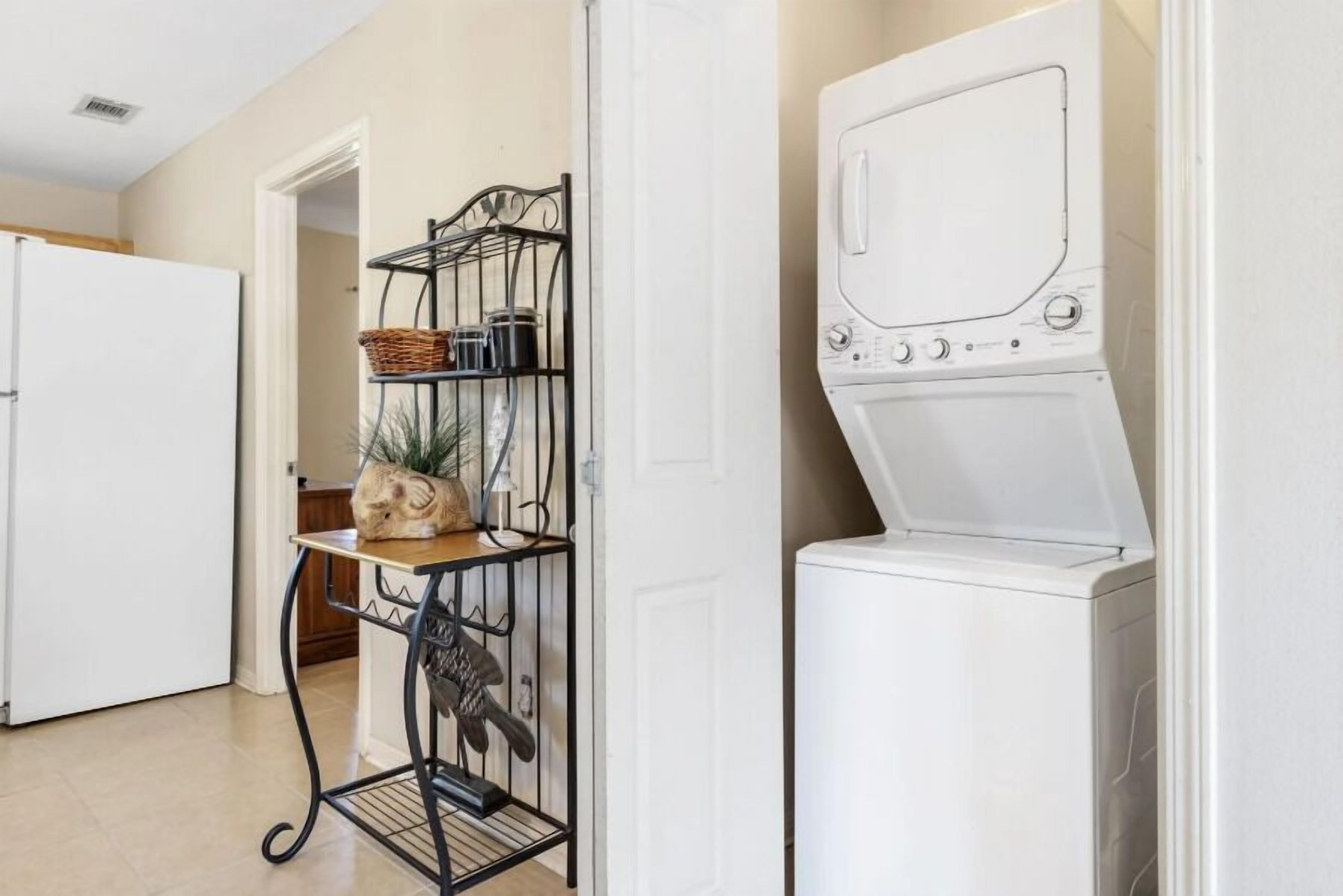 A laundry room with a white washing machine, a metal drying rack, and a shelf with baskets and jars.