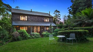 A stone house with a two-story structure, a covered porch, and a well-maintained garden with a lawn and two metal chairs. - StayAU Luxury Candlewick Cottage in Dandenong (Mount Dandenong)
