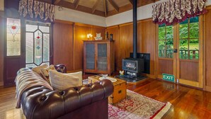 A cozy living room with a leather sofa, a fireplace, and a view of greenery through large windows.