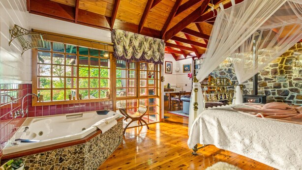 A rustic bedroom with a stone wall, wooden beams, a large window, a bathtub, and a bed with a canopy. - StayAU Owls Croft Picturesque Cottage Valley View (Mount Dandenong)