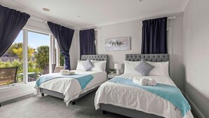 A hotel room with two beds, a large window with curtains, a view of greenery outside, and a painting on the wall.