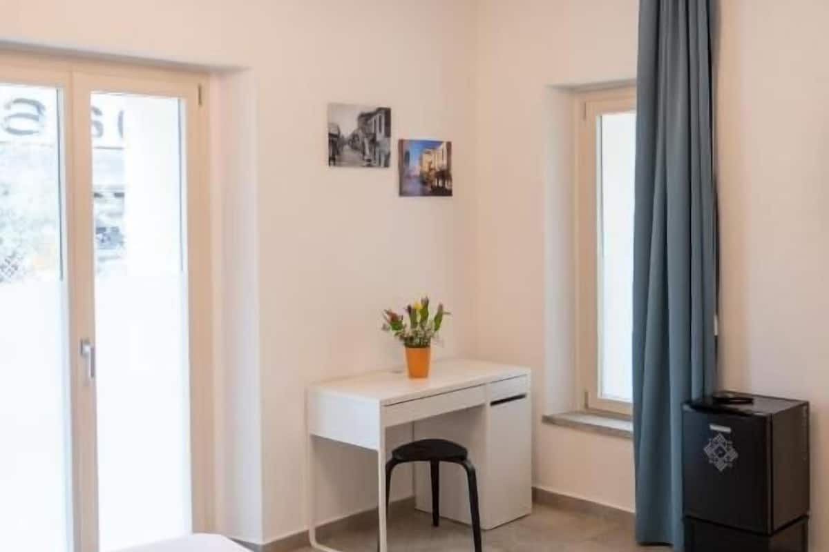 A bedroom with a bed, a desk, a chair, a small table, a potted plant, and a window with curtains.