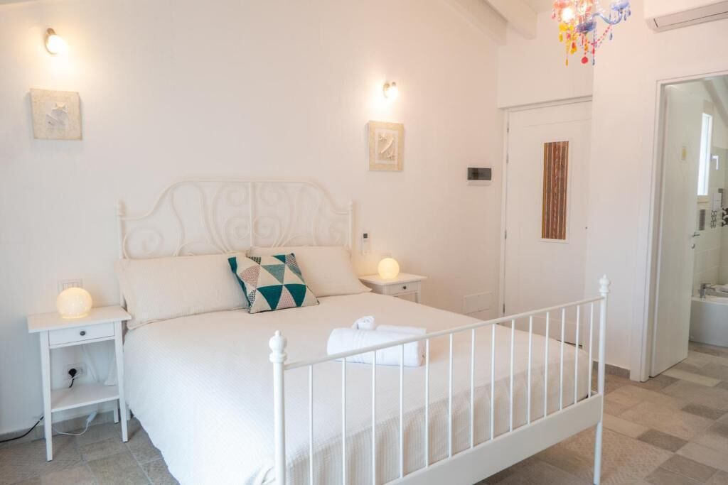 A white four-poster bed with a quilted bedspread and a single decorative pillow. There are two bedside tables with lamps, and a door leading to a bathroom.
