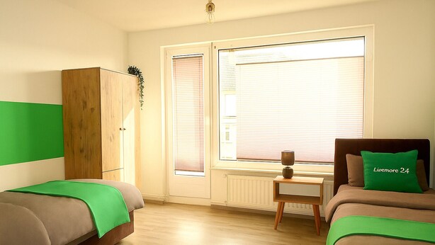 A room with two beds, a wooden wardrobe, a small table with a lamp, and a window with blinds. - Livemore24 - Apartment 2 in Duisburg 24/7 check-in (Duisburg)