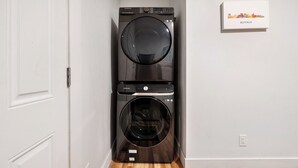 A stacked washer and dryer unit in a hallway with a white door on the left and a framed picture on the wall to the right.