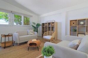 Un salon avec un canapé blanc, une table basse en osier, un fauteuil en osier et une bibliothèque portant l’inscription « MAISON DE VACANCES ».