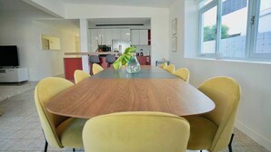 A modern dining area with a wooden table and yellow chairs, a kitchen in the background, and a window with a view. - Villa Coco: Private Pool | 5 Min Walk to Beach (San Juan)