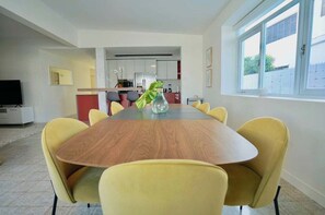 A modern dining area with a wooden table and yellow chairs, a kitchen in the background, and a window with a view. - Villa Coco: Private Pool | 5 Min Walk to Beach (San Juan)