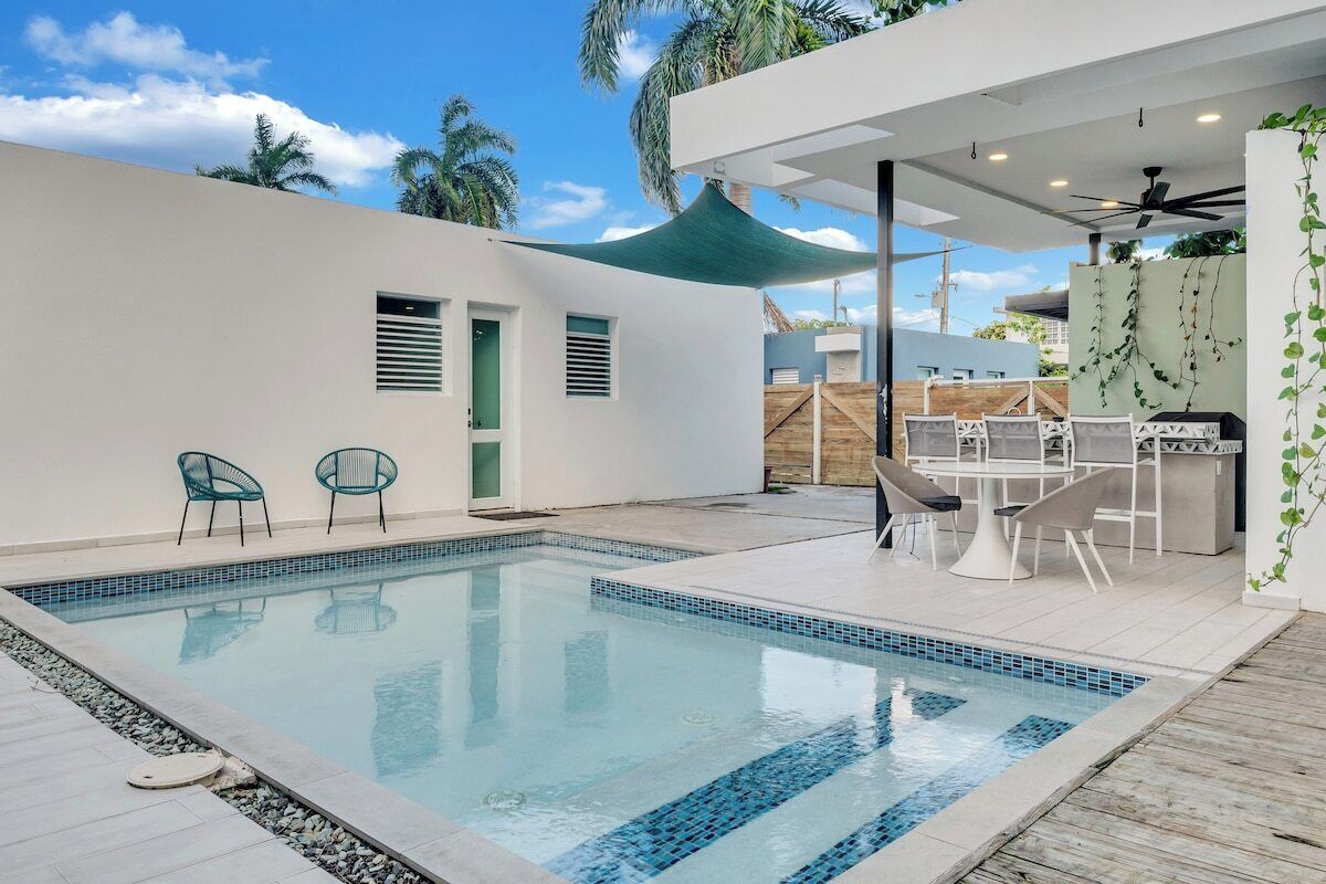 A swimming pool with a tiled edge, surrounded by a patio area with chairs and a table.