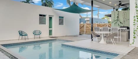 A swimming pool with a tiled edge, surrounded by a patio area with chairs and a table.