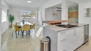 A modern kitchen with a central island, a dining area with yellow chairs, and a view through an open door. - Villa Coco: Private Pool | 5 Min Walk to Beach (San Juan)