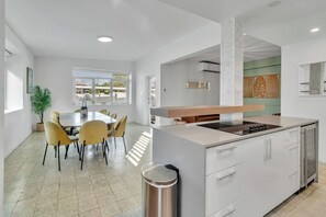 A modern kitchen with a central island, a dining area with yellow chairs, and a view through an open door. - Villa Coco: Private Pool | 5 Min Walk to Beach (San Juan)