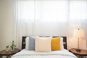 Een bed met een witte dekbedovertrek, vier kussens in verschillende texturen, een bedlampje en een bijzettafeltje met een plant en een mok.