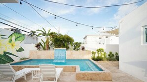 A swimming pool with a tiled edge, surrounded by white walls and a mural. There are lounge chairs and a small table.