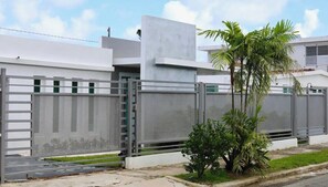 Zona residencial amurallada con una casa moderna, una reja metálica y una pequeña palmera.