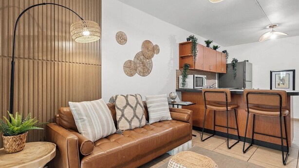 A modern living room with a brown leather sofa, two bar stools, a round coffee table, and a woven ottoman. - Ocean View Rooftop | Rattan Retreat – 1BR (San Juan)