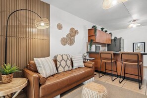 A modern living room with a brown leather sofa, two bar stools, a round coffee table, and a woven ottoman.