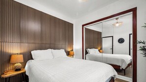 A bedroom with a large bed, a bedside table with a lamp, a mirror, and a clock on the wall. - Ocean View Rooftop | Rattan Retreat – 1BR (San Juan)