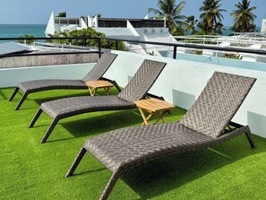 Sillones de exterior sobre césped artificial con vista al océano y edificios al fondo.