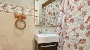 A bathroom with a toilet, a sink, and a shower curtain with a sea shell pattern. - Ocean View Rooftop | Rattan Retreat – 1BR (San Juan)