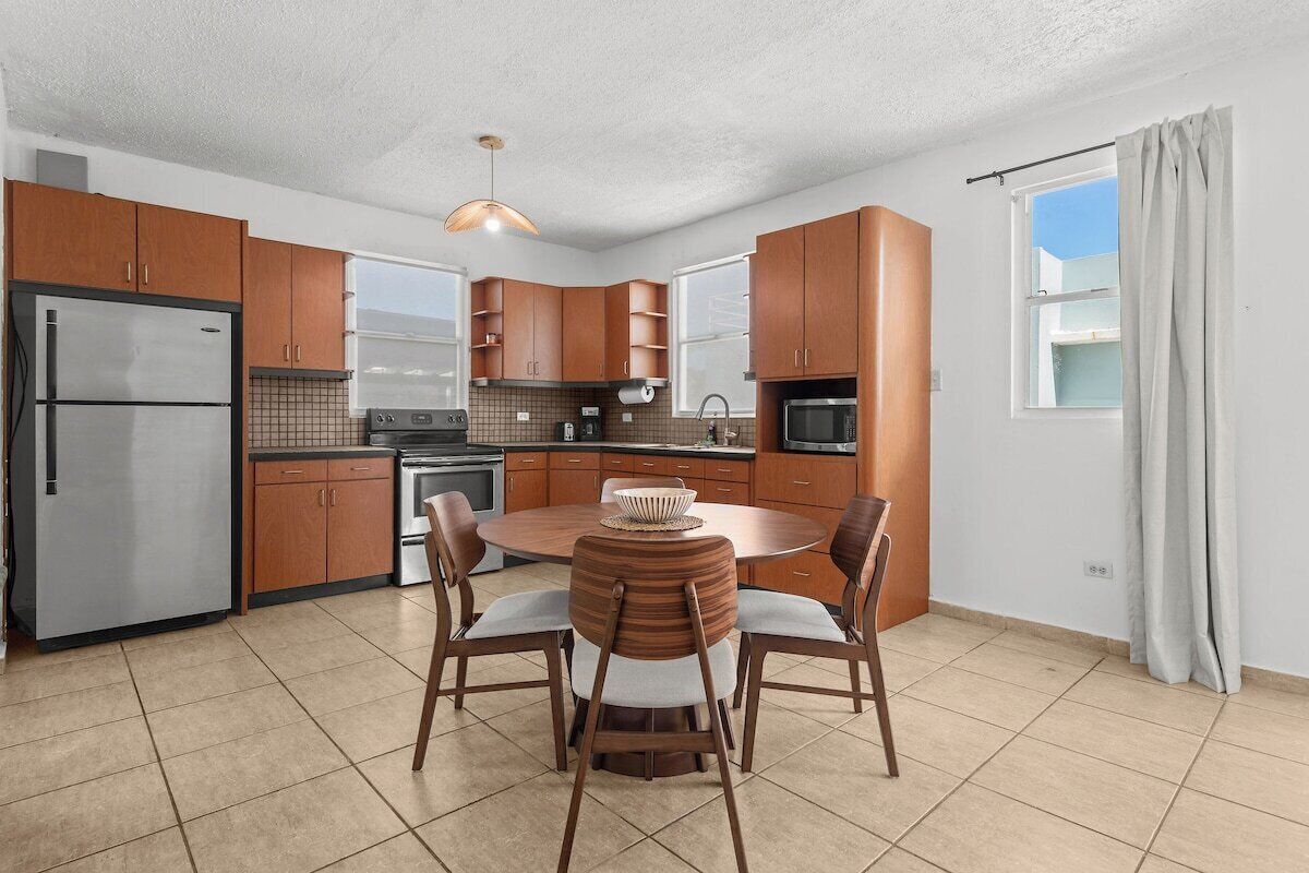 A modern kitchen with wooden cabinets, a stainless steel refrigerator, a round dining table with chairs, and a window with curtains.