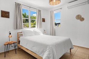 Un dormitorio con una cama, dos ventanas, un aire acondicionado instalado en la pared y una lámpara colgante.